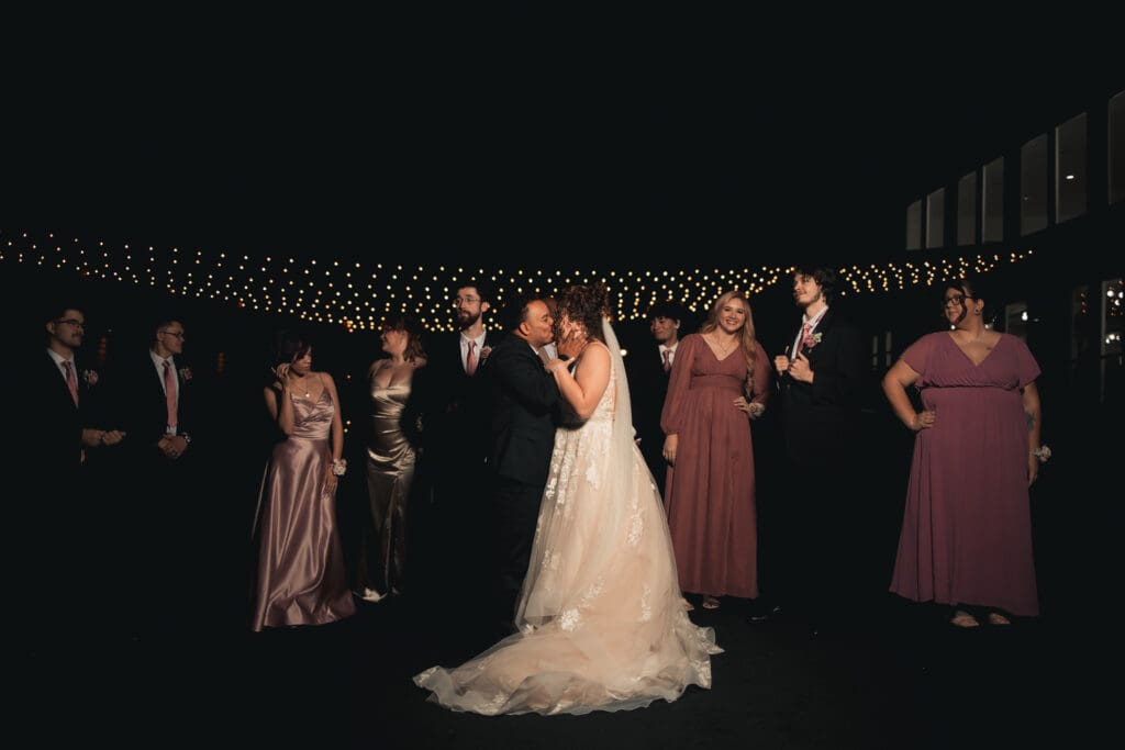 Night time photo of a Springfield area wedding with bright flash. Bride and Groom embrace each other tightly while kissing. Their bridal party stands around them slightly spaced out not looking at the camera to appear cool.
