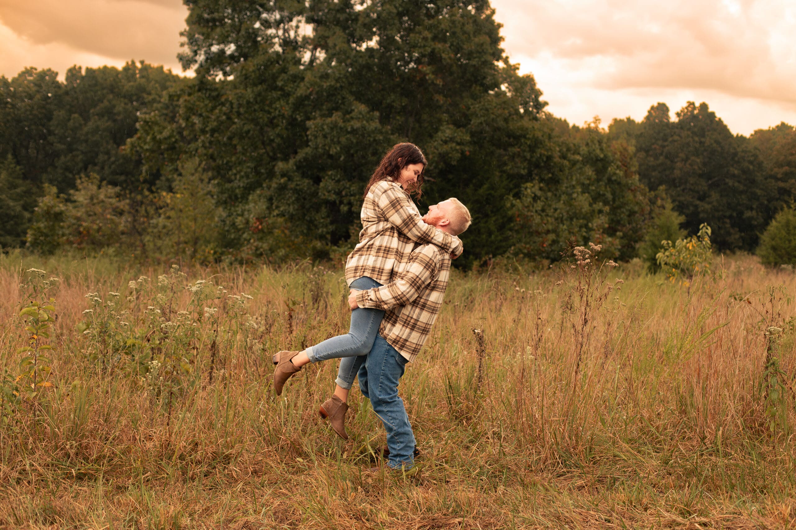 Man and woman in a field wearing flannel shirts during their engagement session. he lifts her by her butt to spin her in the air. They smile at each other lovingly.
