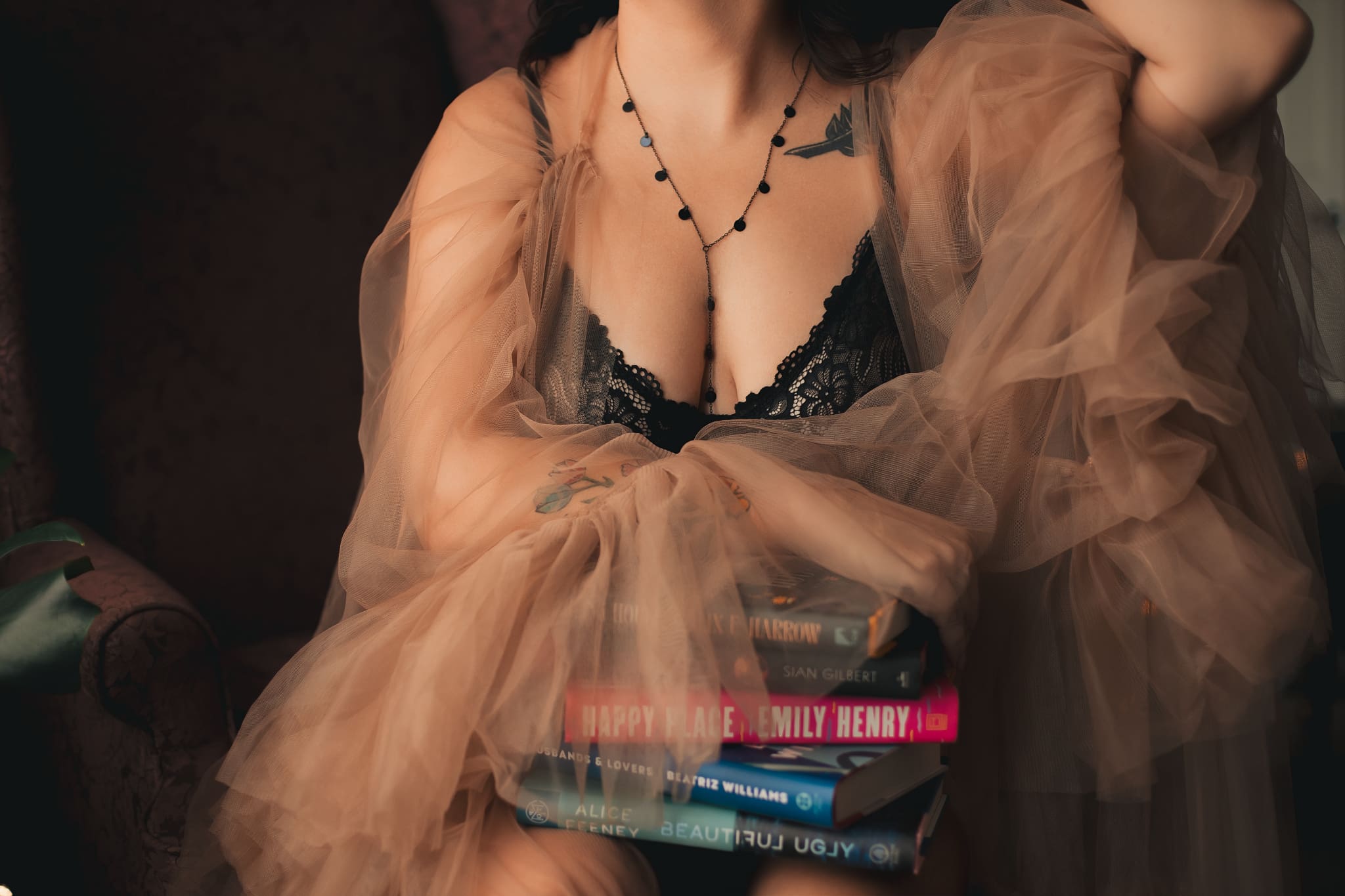 Woman holds books on lap just underneath her breasts. She's wearing a lace black bodysuit that plunges down to her sternum and a nude tulle robe.
