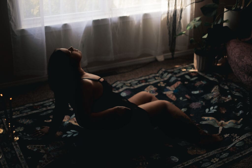 Woman lays on the ground. She's wearing a black sheer dress with slits all the way up to her waist on both sides of her legs. She grabs her underwear and lifts them to the ceiling. Her boudoir shoot is in a dark room with dark accents